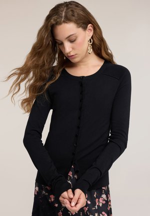 Giovane donna con lunghi capelli ondulati che indossa orecchini d’oro, un cardigan nero con bottoni e una gonna a fiori, guarda verso il basso con le mani giunte.