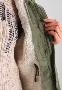 Olivgrüne Jacke mit Fleecefutter, ausgestattet mit Metallschnappverschlüssen und einem Kordelzugsdetail. Beiger Strickpullover zeigt ein strukturiertes Muster.