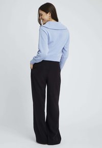 Pull en tricot bleu clair avec un large col, des poignets et un ourlet côtelés ; associé à un pantalon noir à jambes larges, les mains dans les poches.
