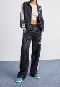 Svart satin joggingdräkt med vita ränder på ärmarna och sidorna, med blommig broderi på jackan och byxorna. Ljust färgade sneakers.