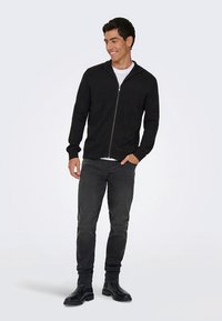 Černá bunda na zip s texturovaným pleteným vzorem, v kombinaci s bílým tričkem, tmavými džínami a černými kotníkovými botami. Model stojí na jednobarevném pozadí.