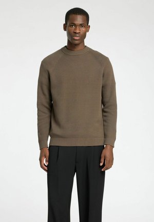 Pull en maille côtelée marron avec un col rond et des manches raglan, porté avec un pantalon noir, modèle se tenant devant un fond neutre.