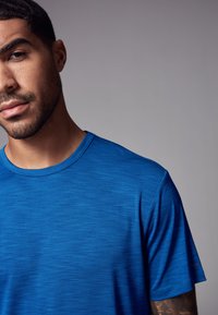 Giovane uomo con capelli corti scuri e barba, indossa una maglietta blu con scollo rotondo, contro uno sfondo grigio uniforme, rivolto a metà verso avanti.