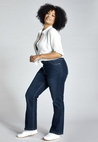 Vaqueros de campana en azul oscuro, con una cintura de altura media, complementados con una camisa blanca de botones y zapatillas blancas. Textura suave.