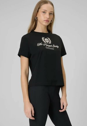 Jonge vrouw met lang blond haar die een zwart T-shirt draagt met het logo van "The Racquet Society" en een zwarte broek tegen een grijze achtergrond.