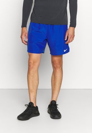 Uomo con pantaloncini Nike blu brillanti, scarpe da corsa nere e un top nero a maniche lunghe, in piedi contro uno sfondo bianco uniforme.