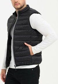 Gilet matelassé noir avec couture horizontale, col haut et fermeture éclair à l'avant. Porté par-dessus une chemise blanche à manches longues, avec une coupe ajustée.