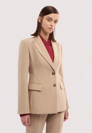 Femme portant un blazer beige cintré et un pantalon assorti sur une chemise à col rouge foncé, debout devant un fond clair uni.