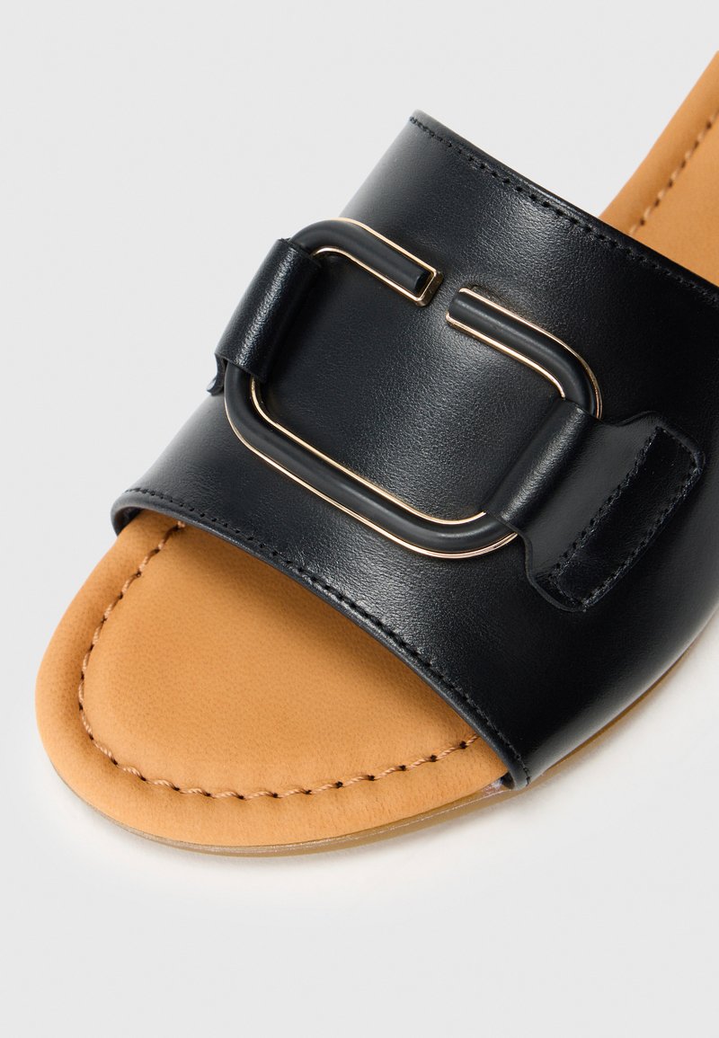 Schwarze Leder-Slipper mit glänzender Oberfläche, versehen mit einer rechteckigen goldfarbenen Schnallenakzent und einer glatten beigefarbenen Innensohle.