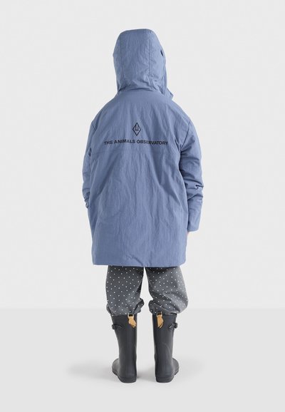 Blaue wasserdichte Jacke mit Kapuze, mit einem Logo auf dem Rücken, kombiniert mit grauen gepunkteten Hosen und schwarzen Gummistiefeln.