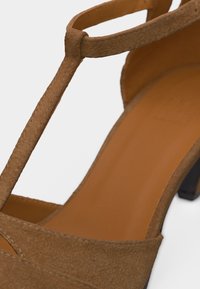 Καφέ σουέντ πέδιλο με λουράκι σε σχήμα Τ, ψηλό τακούνι, με λείο ταμπά πάτο και μαύρο τακούνι, σε λευκό φόντο.