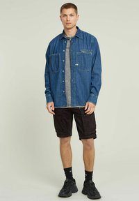 Camicia in denim di colore blu scuro con due tasche frontali e una maglietta grigia a contrasto sotto. Abbinata a pantaloncini cargo neri e sneakers nere.
