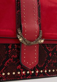 Bolso rojo con texturas de ante y charol. Presenta un estampado de serpiente, tachuelas doradas y un acento de cadena dorada en la correa. Elementos de diseño distintivos.