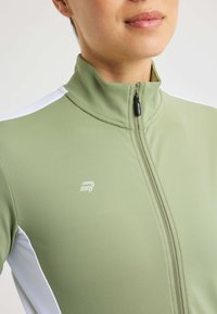Ljusgrön zip-jacka med hög krage, vita paneldetaljer och en liten logotyp på bröstet. Slät, elastisk tygstruktur.