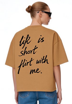 Femme avec un chignon et des lunettes de soleil porte une chemise marron surdimensionnée avec "life is short flirt with me." écrit en noir dans le dos.
