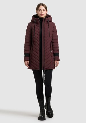 Cappotto imbottito bordeaux con cappuccio, design a righe verticali, chiusura con zip e dettagli neri. Indossato sopra leggings neri e stivaletti.