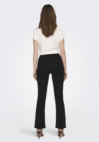 Witte top met korte mouwen en kantafwerking, gecombineerd met nauwsluitende zwarte flared pants en zwarte hakken. Achteraanzicht, minimale achtergrond.