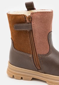 Bottines marron et beige avec une fermeture éclair et une doublure en peluche. Présente un mélange de matériaux lisses et texturés avec une semelle robuste.