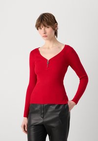 Haut long à manches ribbé rouge avec un décolleté en V profond et un zip au niveau du col, associé à un pantalon en cuir noir.