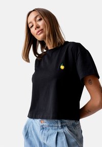 Czarna krótka koszulka wykonana z bawełny, z małym haftem żółtej cytryny na piersi; modelka nosi jasnoniebieskie spodnie z wysokim stanem.
