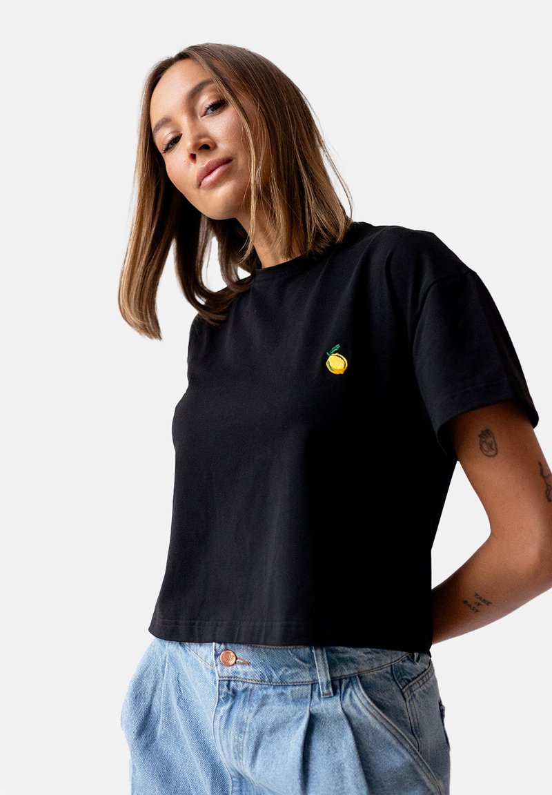 Czarna krótka koszulka wykonana z bawełny, z małym haftem żółtej cytryny na piersi; modelka nosi jasnoniebieskie spodnie z wysokim stanem.
