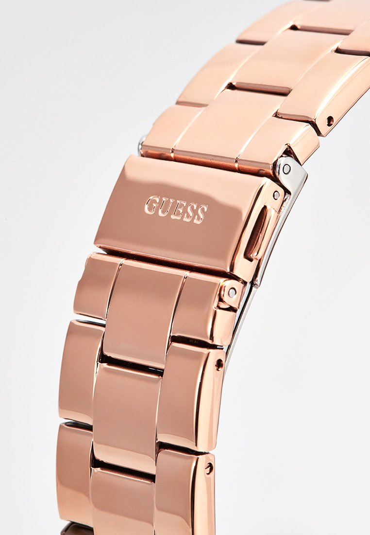 Pulsera de reloj de acero inoxidable en color rosa dorado con eslabones rectangulares y acabado pulido. Presenta el logotipo grabado "GUESS" en el cierre.