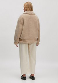 Beige schapenvachtjack met een hoge kraag, oversized vorm en een ceintuur in de taille, gecombineerd met crème brede broekspijpen en zwarte schoenen.