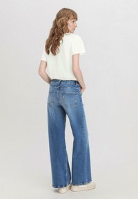 Siniset leveälahkeiset farkut haalistuneella pesulla, korkea vyötärö ja takataskut, yhdistetty valkoiseen lyhythihaiseen t-paitaan ja vaaleisiin kenkiin.