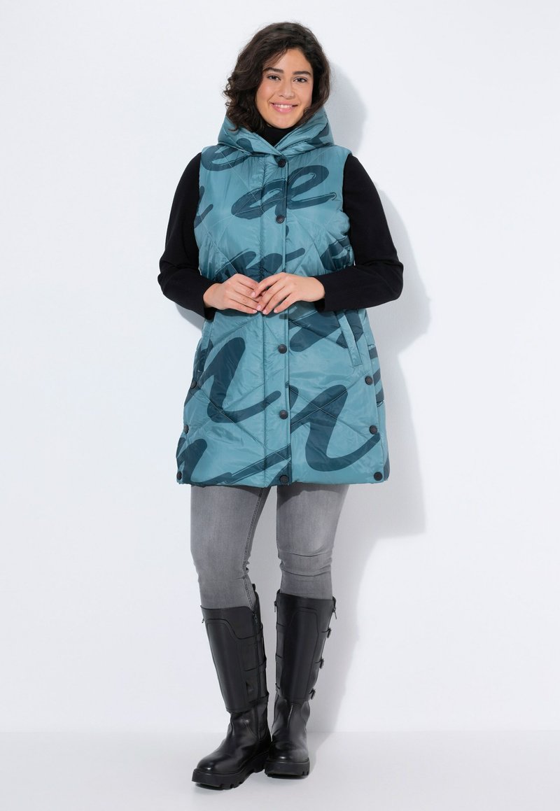 Vadderad teal väst med svart abstrakt mönster, hög krage, knappstängning och sidofickor, bärs över en svart långärmad topp och grå jeans.