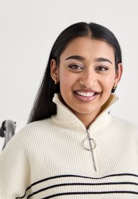 Maglione a coste beige chiaro con colletto con zip, dotato di una pendente a anello argentato e strisce orizzontali blu navy sul petto.