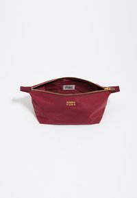 Bolso de cremallera en tonos marrón oscuro, confeccionado con un tejido texturizado y con un acento de logo dorado y herrajes dorados en la cremallera. Interior forrado con tela en color marrón oscuro.