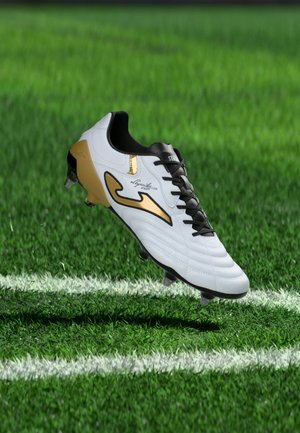 Chaussure de football blanche et dorée avec des lacets noirs flottant au-dessus de l'herbe verte près d'une ligne blanche du terrain.