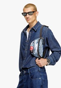 Camicia di jeans con taschino, pantaloni cargo blu e una borsa piccola con design in denim strappato e logo rosso, indossata a tracolla. Occhiali da sole neri.