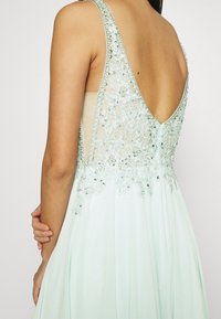 Mátově zelené šaty s nízkým výstřihem na zádech, zdobené jemnou výšivkou a korálkovými detaily. Plynulá látka padá až na podlahu, čímž vytváří jemnou siluetu.