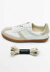 Valkoinen nahkakenkä, jossa on beige mokkanahka koristeita ja ruskea kumipohja. Mukana on matching-beige nauhat, joissa on musta bändilappu.