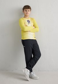 Hosszú ujjú, sárga sportos póló logóval, fekete nadrág és világosszürke sportcipő; egy texturált világosszürke falnak támaszkodva áll.