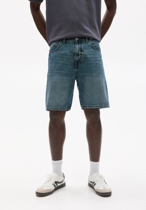 Homme portant un short en jean bleu mi-long, des chaussettes blanches, des baskets blanches et grises, et un t-shirt gris foncé sur fond blanc.