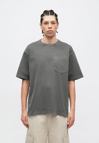 Szürke, túlméretezett pamut póló kerek nyakkal és rövid ujjakkal. Mellkasi zsebbel és látható varrási részletekkel rendelkezik.