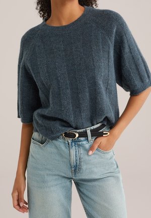 Vrouw draagt een getextureerde donkerblauwe trui met korte mouwen, ingestopt in lichtblauwe spijkerbroek met een zwarte riem, hand in de zak.