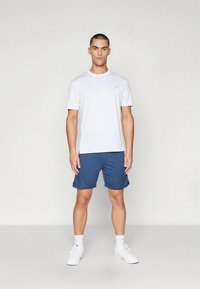 Vit t-shirt med korta ärmar och en minimal logotyp, kombinerad med marinblå sportshorts och vita sneakers. Modellen står framåtvänd mot en enfärgad bakgrund.