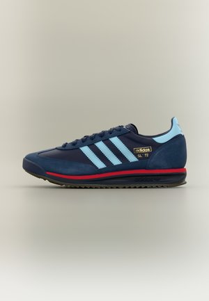 Sneaker in suede e materiale sintetico colore blu navy con tre strisce azzurro chiaro, suola nera e intersuola rossa. Dettaglio logo dorato sul lato. Design classico.