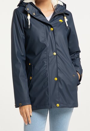Femme portant un imperméable bleu marine avec des boutons jaunes, des cordons blancs avec des perles en bois et un petit logo parapluie jaune sur la poitrine.