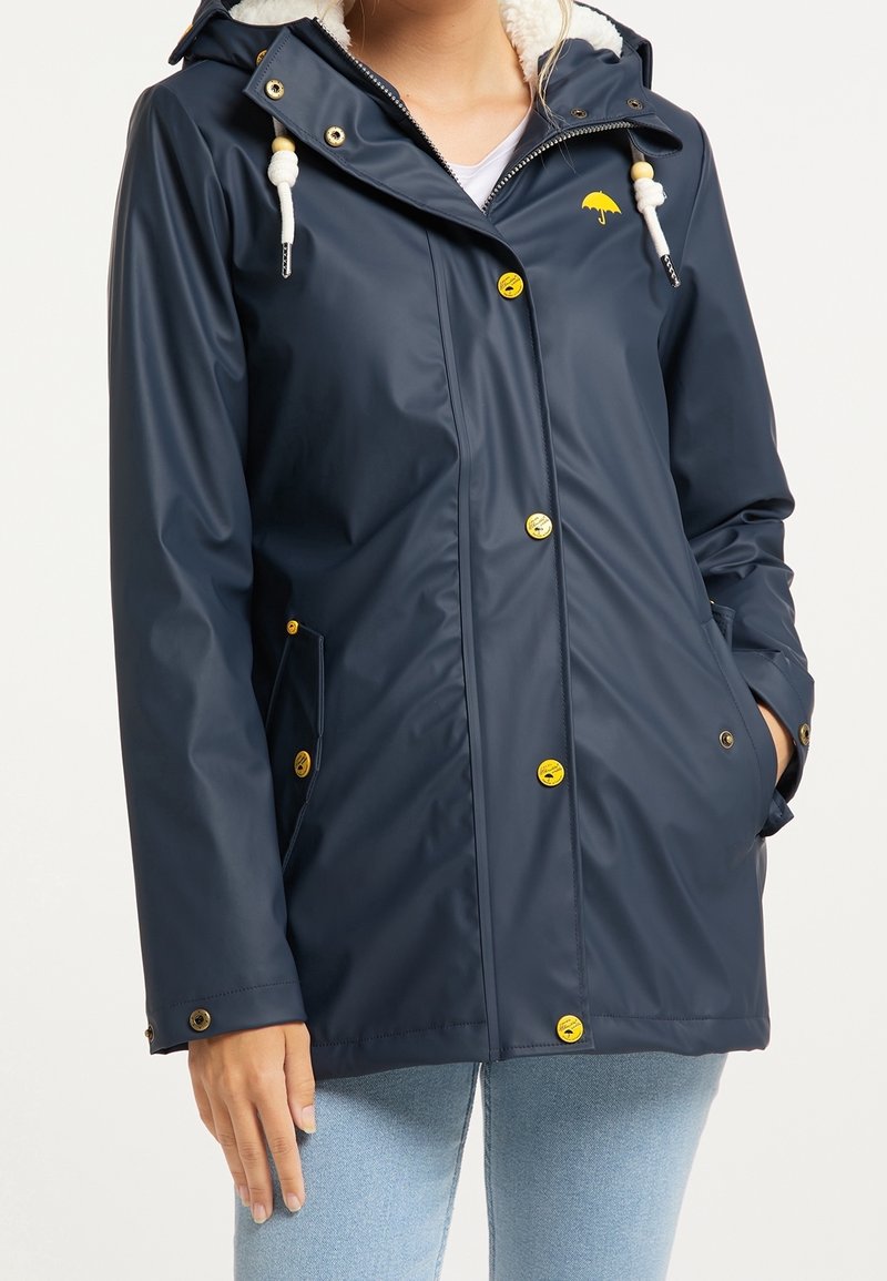 Femme portant un imperméable bleu marine avec des boutons jaunes, des cordons blancs avec des perles en bois et un petit logo parapluie jaune sur la poitrine.