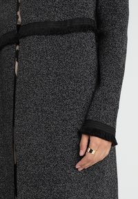 Detailní záběr na černobílý texturovaný kabát s třásněmi na manžetách a pase, který ukazuje ruku nosící široký zlatý prsten.