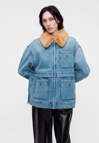 Jeune personne aux cheveux courts et foncés portant une veste en jean bleue avec un col en fourrure beige et un pantalon en cuir noir, face à l’avant, les mains derrière.