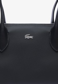 Borsa nera con un'esterno in tessuto texture, caratterizzata da un dettaglio in logo di coccodrillo argentato e due manici robusti. Accenti in pelle liscia.