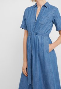 Denimová košilová šaty s krátkými rukávy, límečkem, zúženým pasem, bočními kapsami a délkou po kolena. Světle modrá s jemnou texturou.