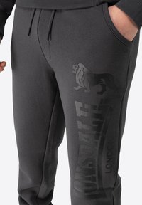 Pantalons de survêtement gris foncé avec une texture lisse, cordon de serrage ajustable, poches latérales et grand logo de lion avec le nom de la marque en impression contrastée.