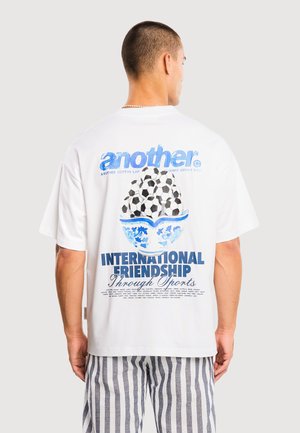 Mężczyzna ubrany w białą koszulkę z niebieskim napisem i grafiką piłki nożnej, w połączeniu z pionowo paskowanymi spodniami, stoi na jednolitym tle.