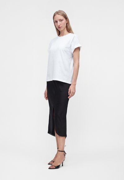 Young woman standing wearing a white short-sleeve top, black midi skirt with front slit, and black ankle-strap high heel sandals.